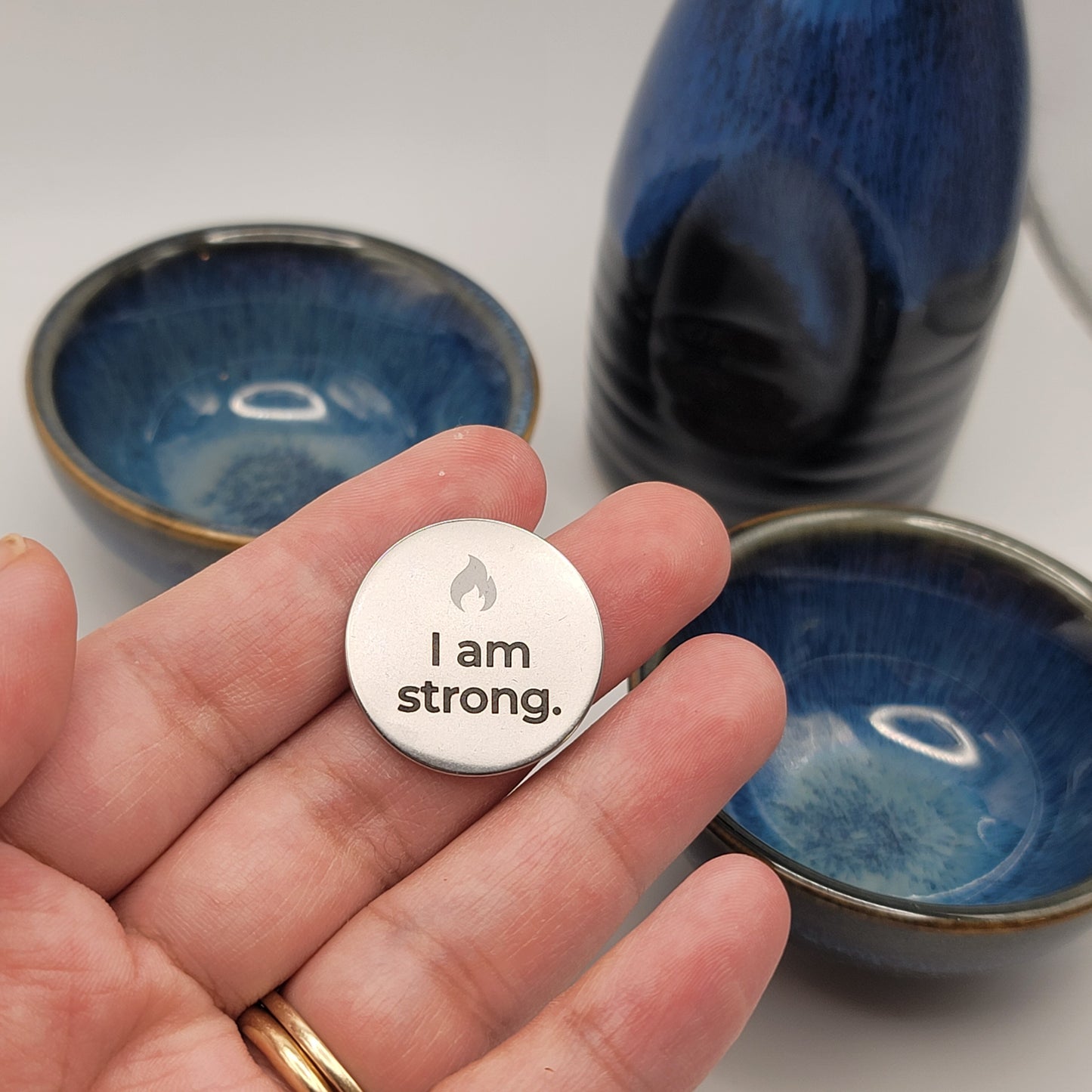 A hand holds a stainless steel affirmation token engraved with the words “I am strong,” with blue ceramic bowls softly blurred in the background, symbolizing calm and mindfulness.