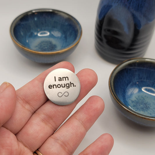 A hand holds a stainless steel affirmation token engraved with the words “I am enough,” with blue ceramic bowls softly blurred in the background, symbolizing calm and mindfulness.