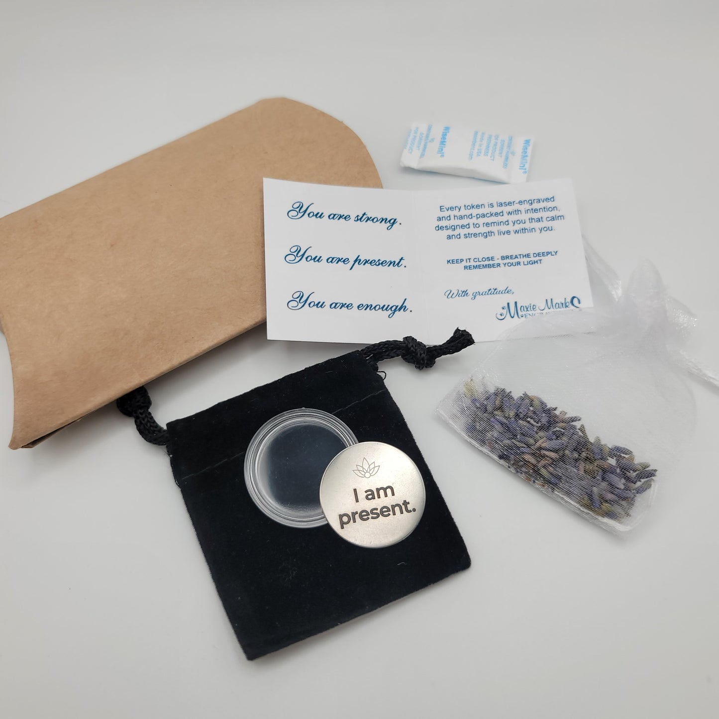 A complete display of the I Am Present affirmation token set, including the engraved token, velvet pouch, acrylic capsule, lavender sachet, and affirmation card with the message “You are strong. You are present. You are enough.”
