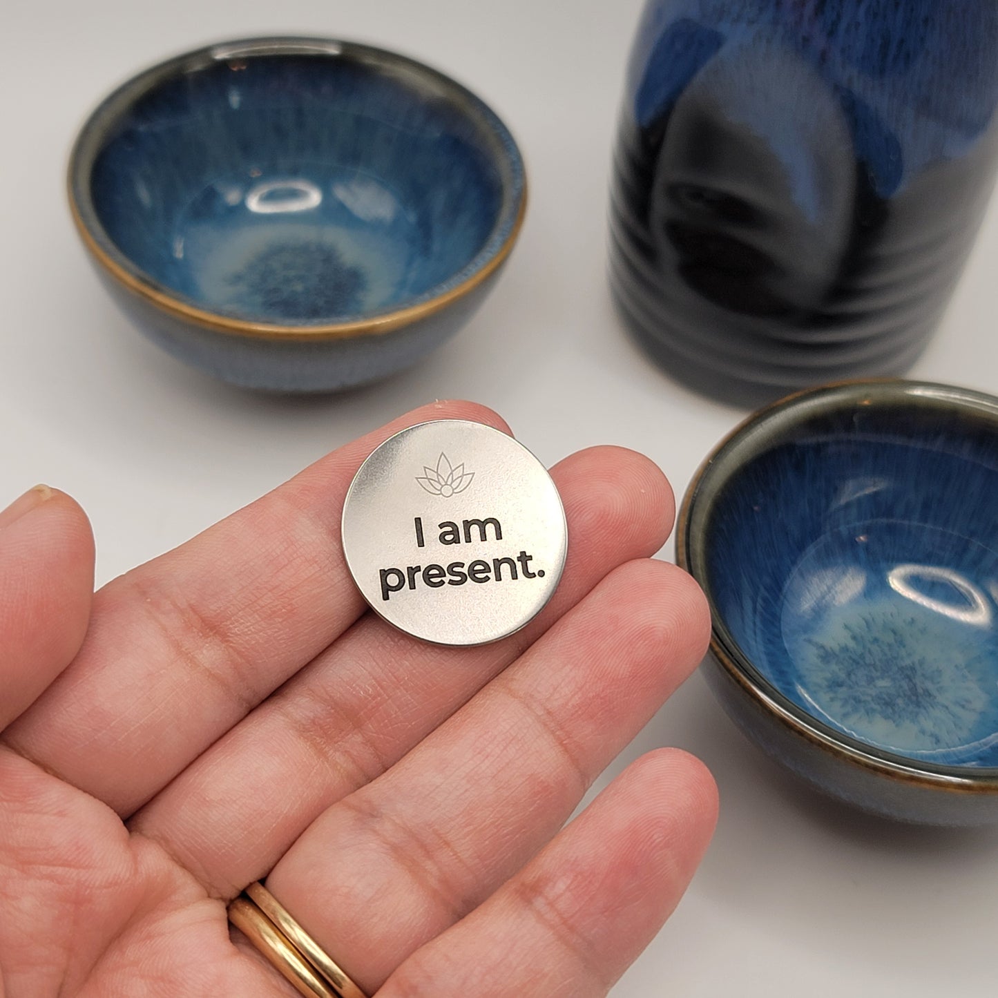 A hand holds a stainless steel affirmation token engraved with the words “I am present,” with blue ceramic bowls softly blurred in the background, symbolizing calm and mindfulness.

