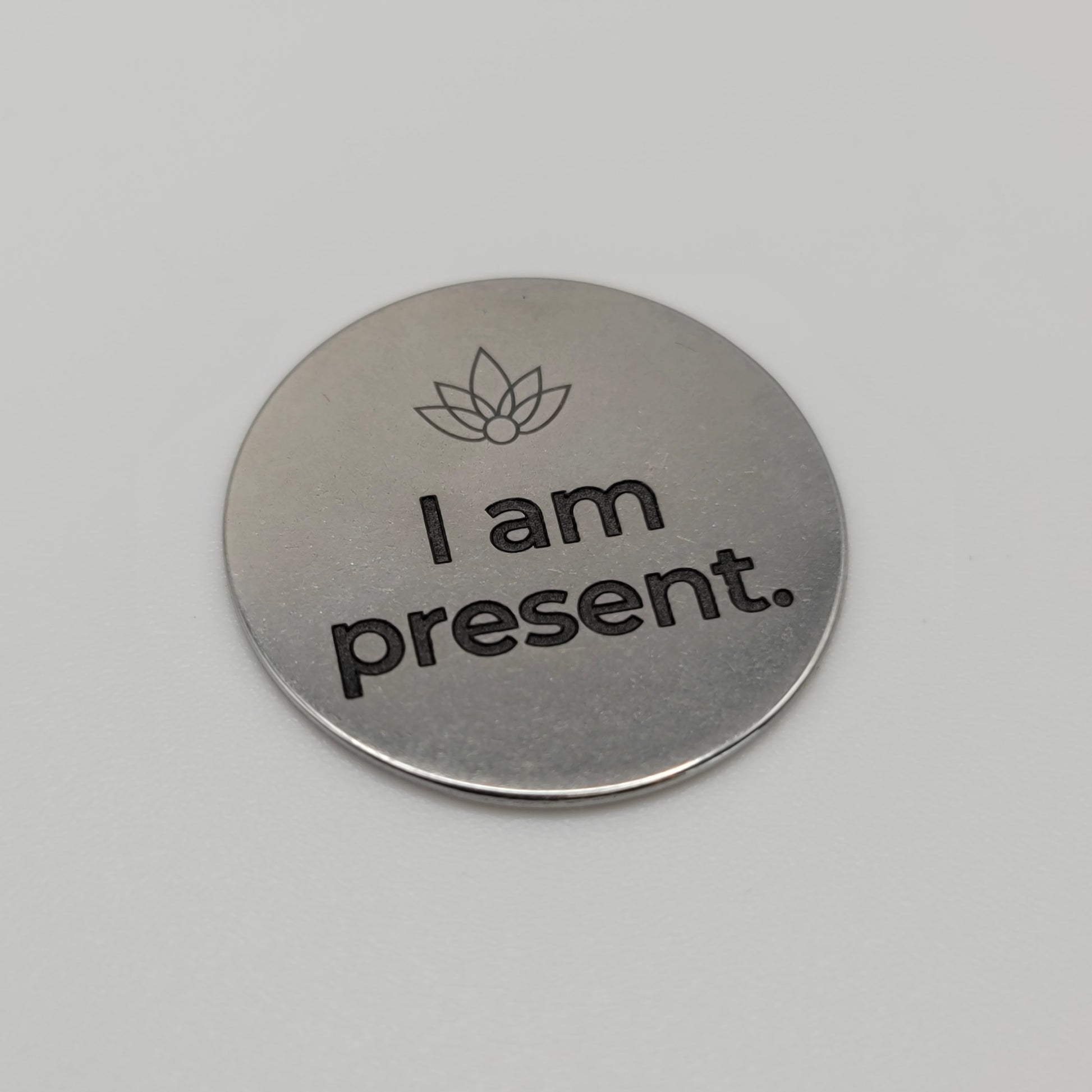 A close-up view of a stainless steel token engraved with a flame symbol and the affirmation “I am present,” placed on a clean white surface to highlight the engraved detail and brushed finish
