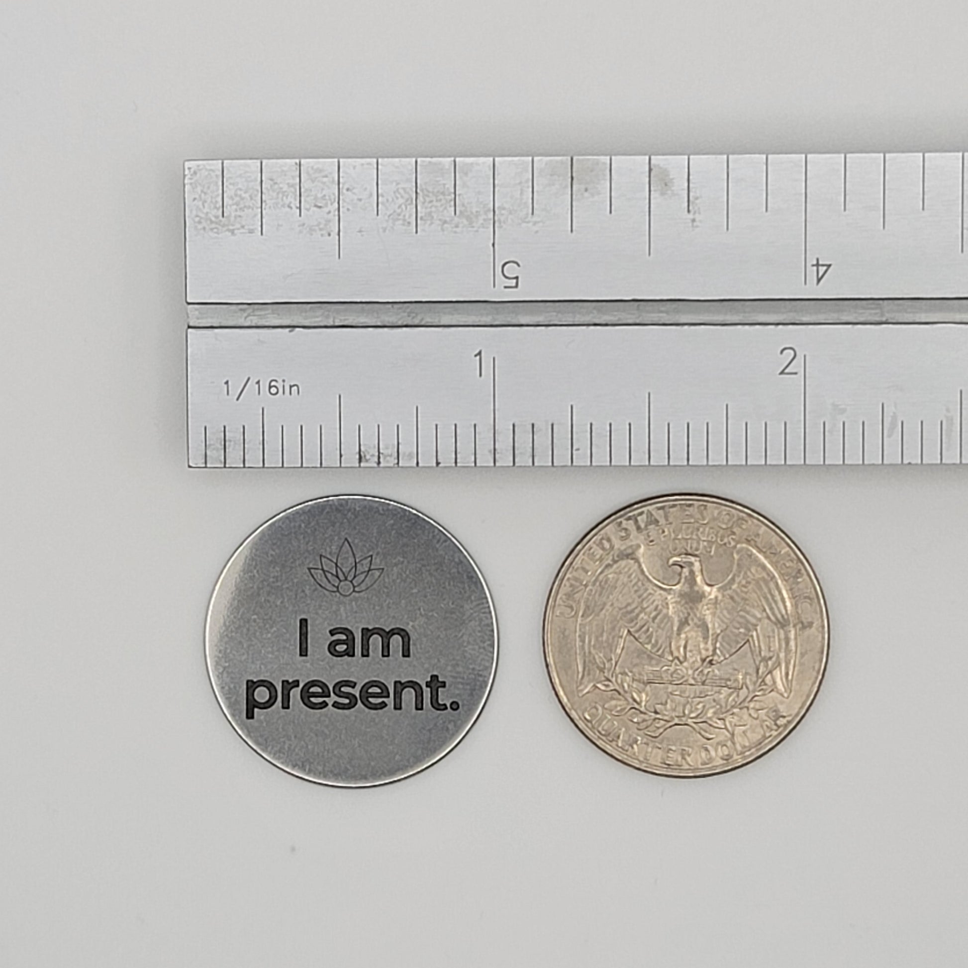 A stainless steel “I am present” affirmation token shown beside a U.S. quarter and a ruler for size reference, illustrating the token’s compact and pocket-friendly dimensions.
