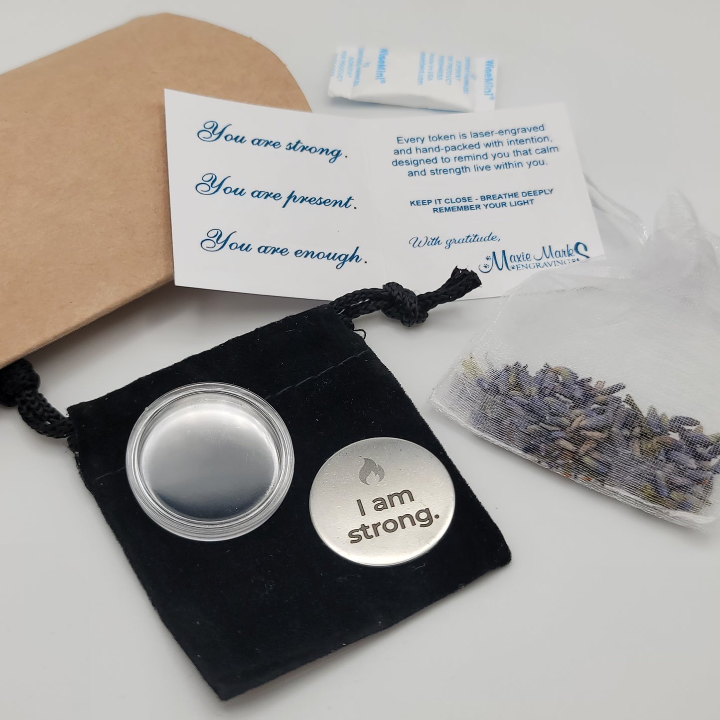 A complete display of the I Am Strong affirmation token set, including the engraved token, velvet pouch, acrylic capsule, lavender sachet, and affirmation card with the message “You are strong. You are present. You are enough.”