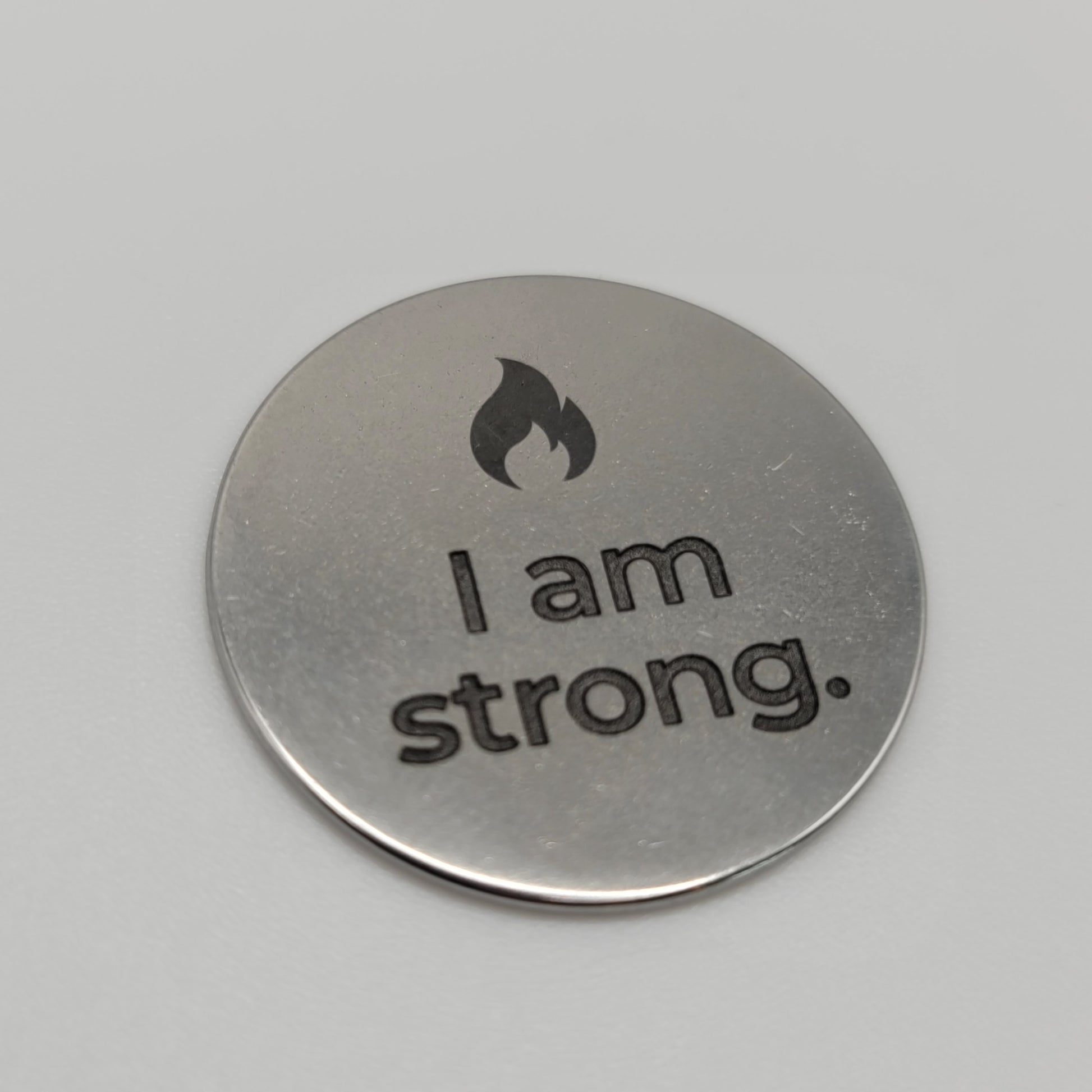 A close-up view of a stainless steel token engraved with a flame symbol and the affirmation “I am strong,” placed on a clean white surface to highlight the engraved detail and brushed finish