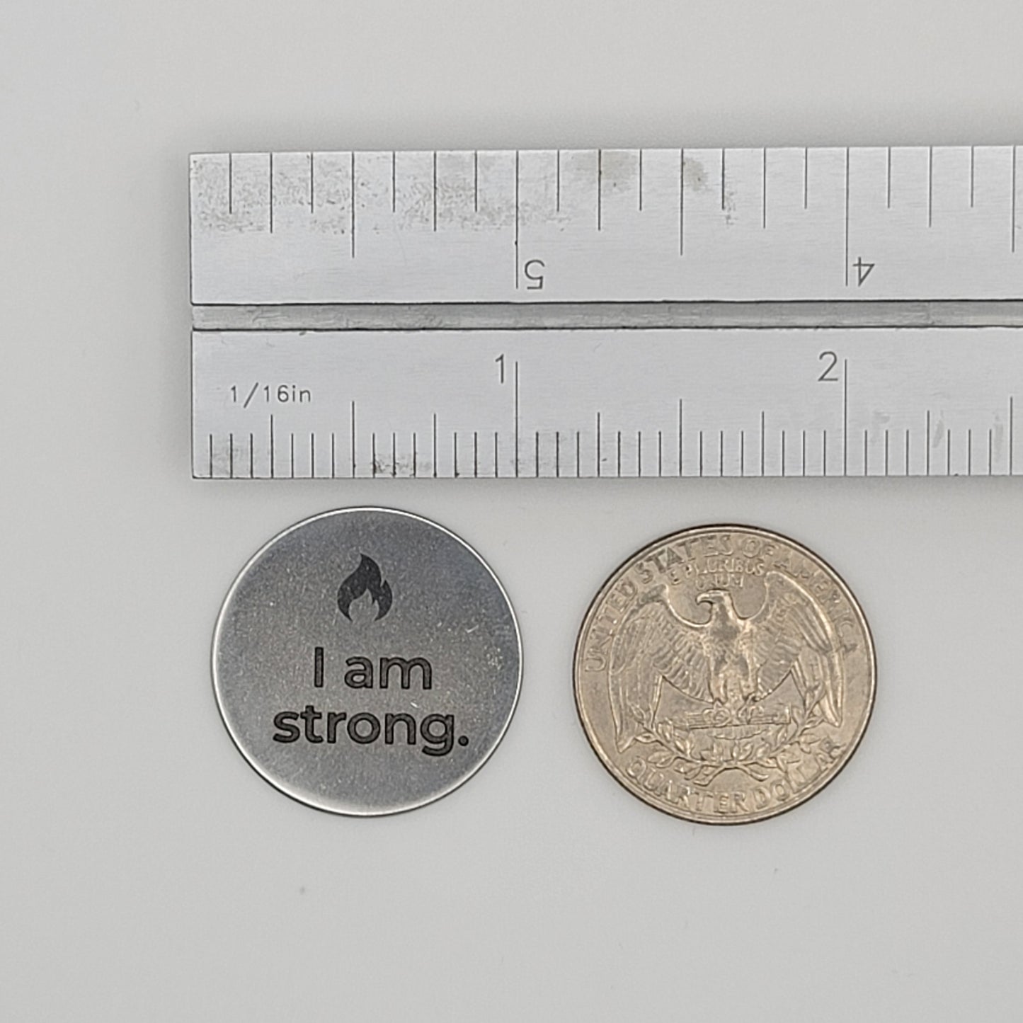 A stainless steel “I am strong” affirmation token shown beside a U.S. quarter and a ruler for size reference, illustrating the token’s compact and pocket-friendly dimensions.