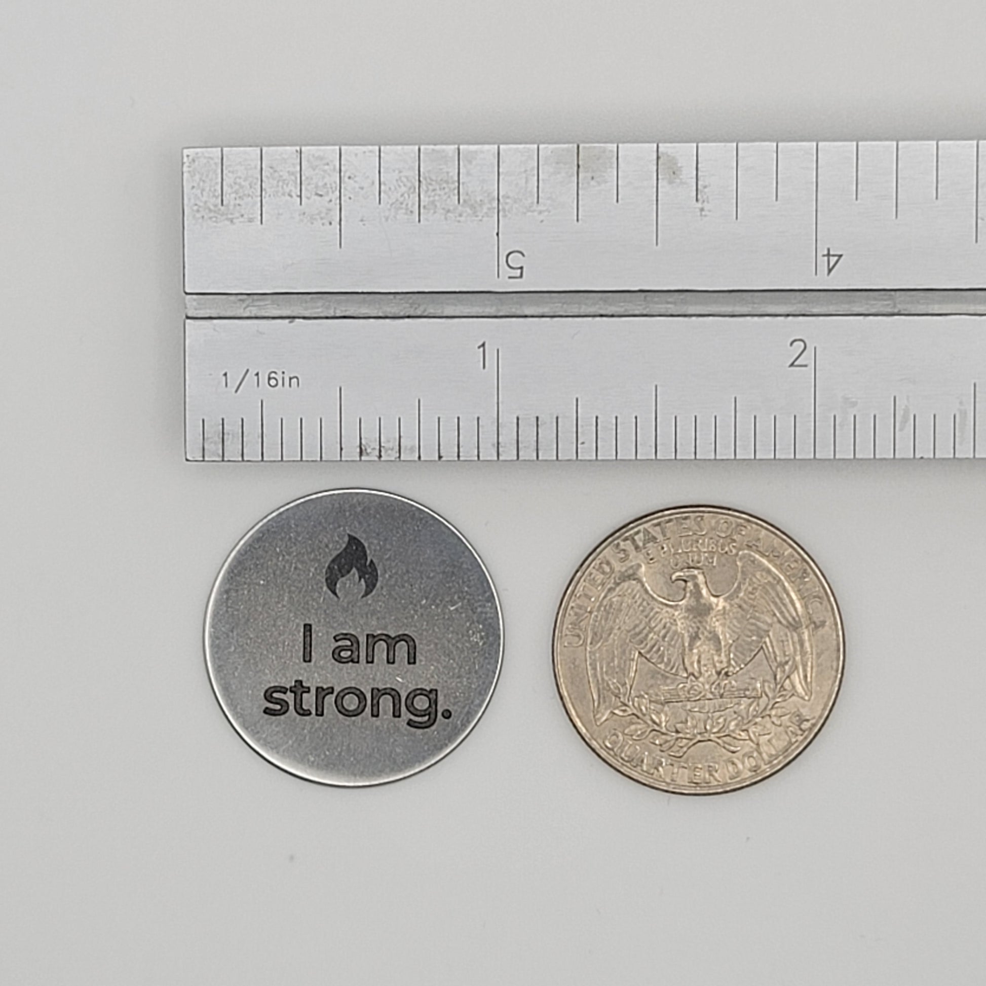A stainless steel “I am strong” affirmation token shown beside a U.S. quarter and a ruler for size reference, illustrating the token’s compact and pocket-friendly dimensions.